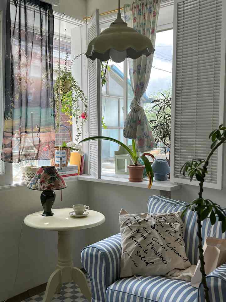 A white-walled veranda space featuring a blue striped armchair, floral table lamp, and various plants creating a natural vintage workspace atmosphere.