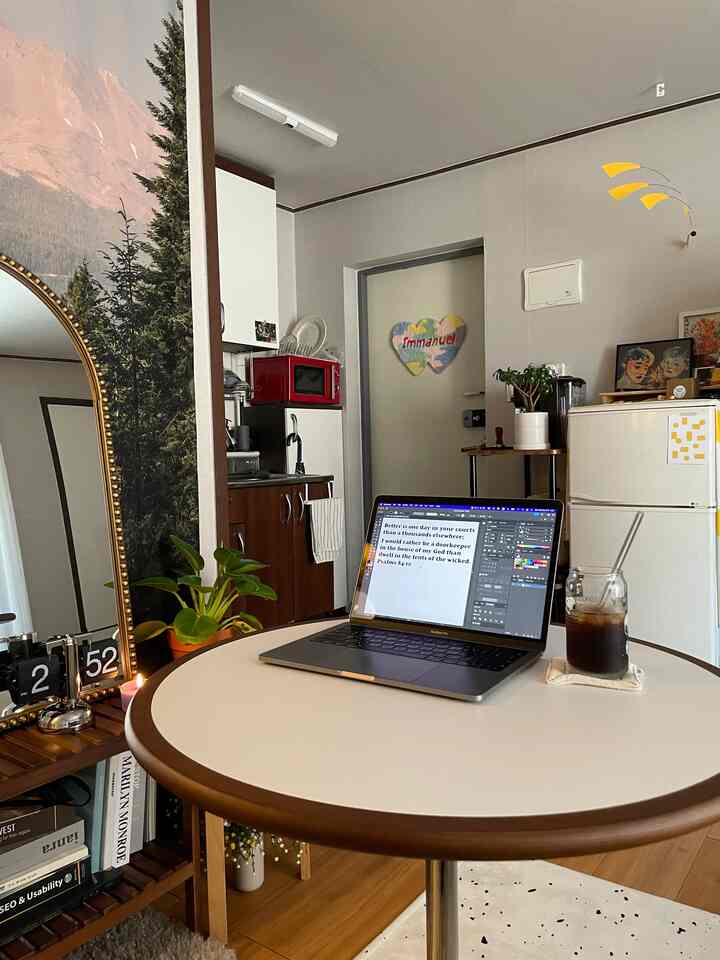 Natural wood-toned living area with visible entrance, featuring a round table with laptop and iced coffee, evoking home cafe and remote work atmosphere