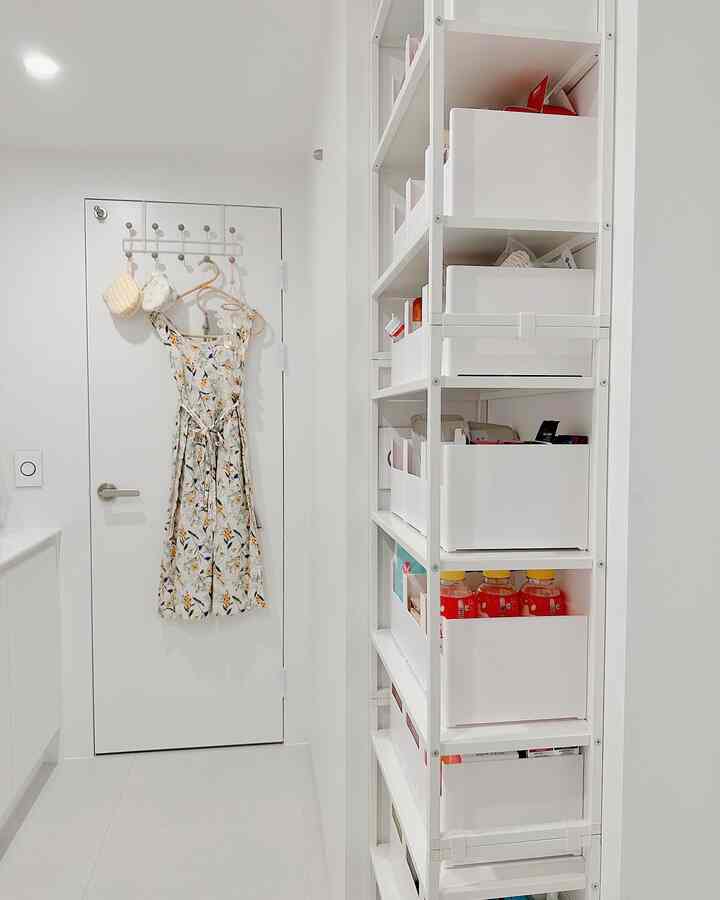 White-toned narrow space featuring a door with coat racks and dress, and a tall white storage cabinet on the right, creating an organized storage area