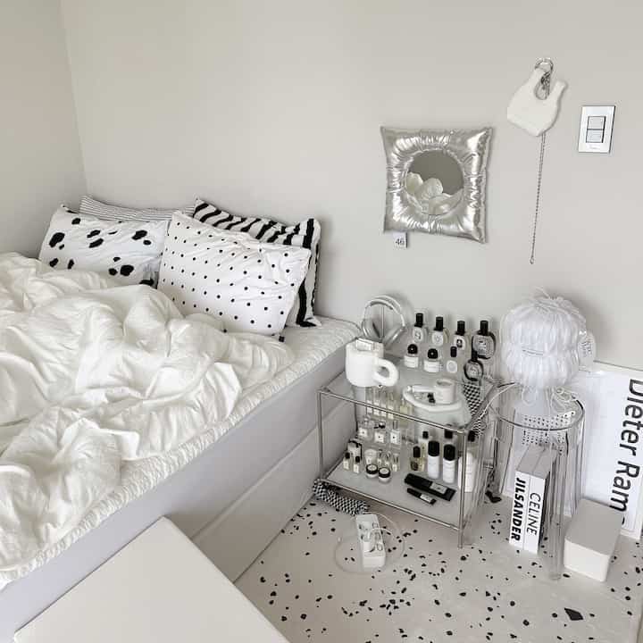 Bright white-toned bedroom featuring white bedding with patterned pillows, transparent stool, and glass shelf, creating a modern minimal atmosphere