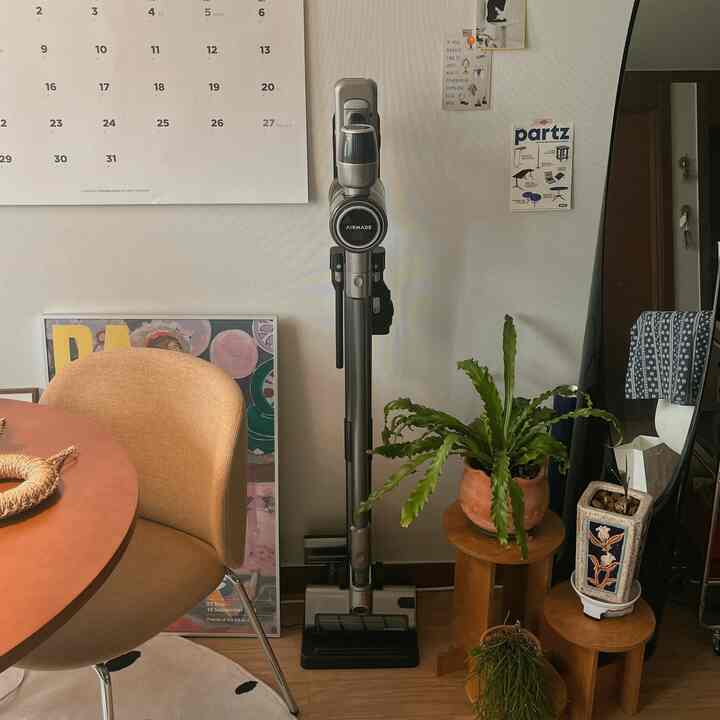 ホワイトとブラウンの色調のリビングルーム、中央にコードレス掃除機があり、周囲に木製スツールと緑の植物が配置された雰囲気のある空間