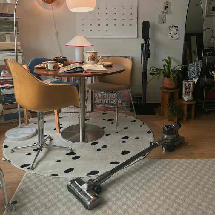 暖かいトーンのワンルームカフェ風空間、丸テーブルと様々な椅子、植物、掃除機が配置された雰囲気のあるリビング