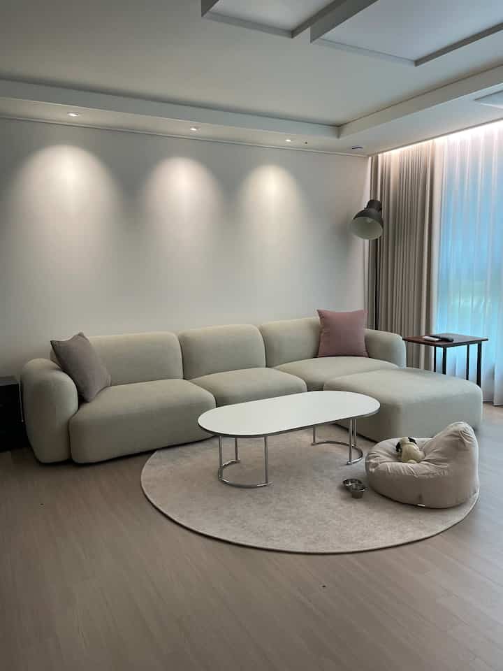 ナチュラルなベージュとホワイトのトーンのリビング、広いソファと丸いラグ、ビーンバッグとフロアランプが配置された落ち着いた空間