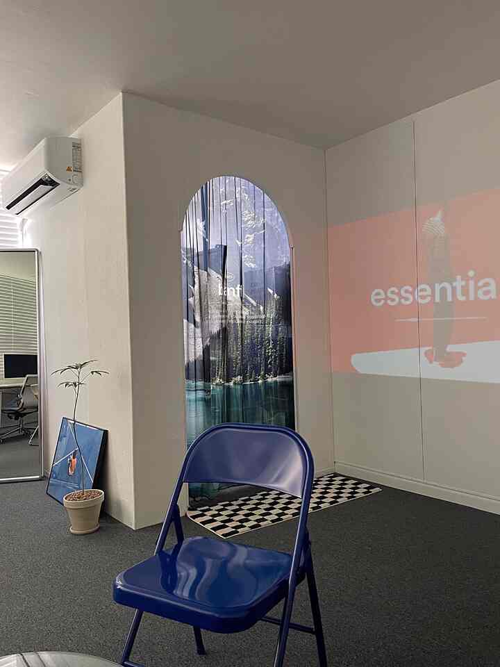 白い壁と天井のミニマルなオフィス空間、中央にブルーの折りたたみ椅子があり、アーチ型の扉には自然の風景のカーテンとチェッカーボード柄のラグが配置されている。