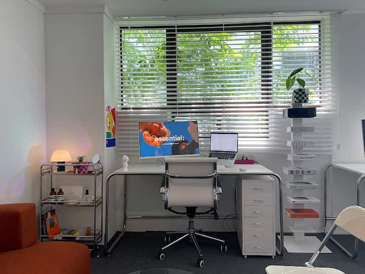 白を基調としたコンパクトなホームオフィス、机とオフィスチェア、本棚、横型ブラインドが配置された落ち着いた空間