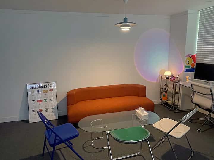 白い壁のミッドセンチュリーモダンなリビング、オレンジのソファとカラフルな折りたたみ椅子、透明なガラスのコーヒーテーブルが配された雰囲気のあるインテリア