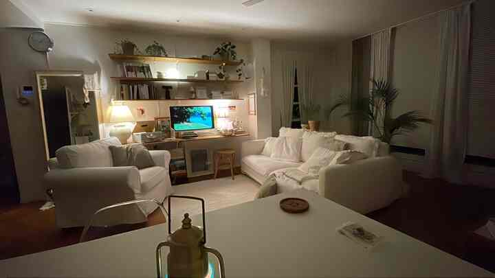 Warm-lit natural tone living room featuring white sofas, curtains, wooden shelves, and a desk creating a cozy atmosphere