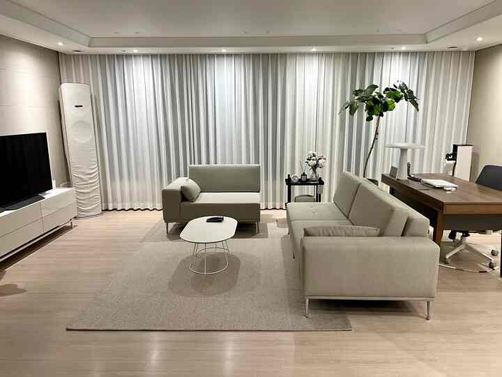 Bright white curtains and wood tone flooring frame a modern living room with sofas and an integrated home office setup