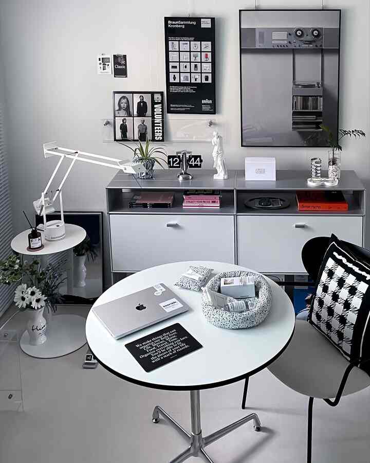 White-toned modern home office featuring a round table, armchair, lighting, and posters creating a clean workspace atmosphere