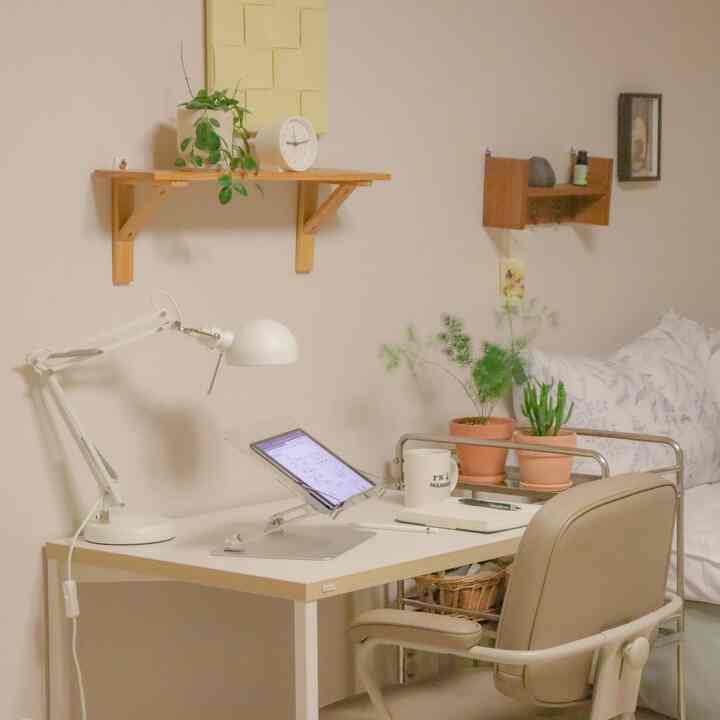 白とベージュの色調が特徴のワンルーム寝室、作業用デスクとオフィスチェア、棚、植物が配置された温かみのある清潔なインテリア