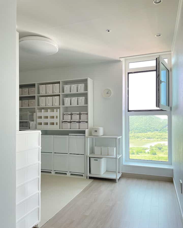 Bright pantry space in white and natural tones, featuring neatly organized storage boxes and a large window with scenic view
