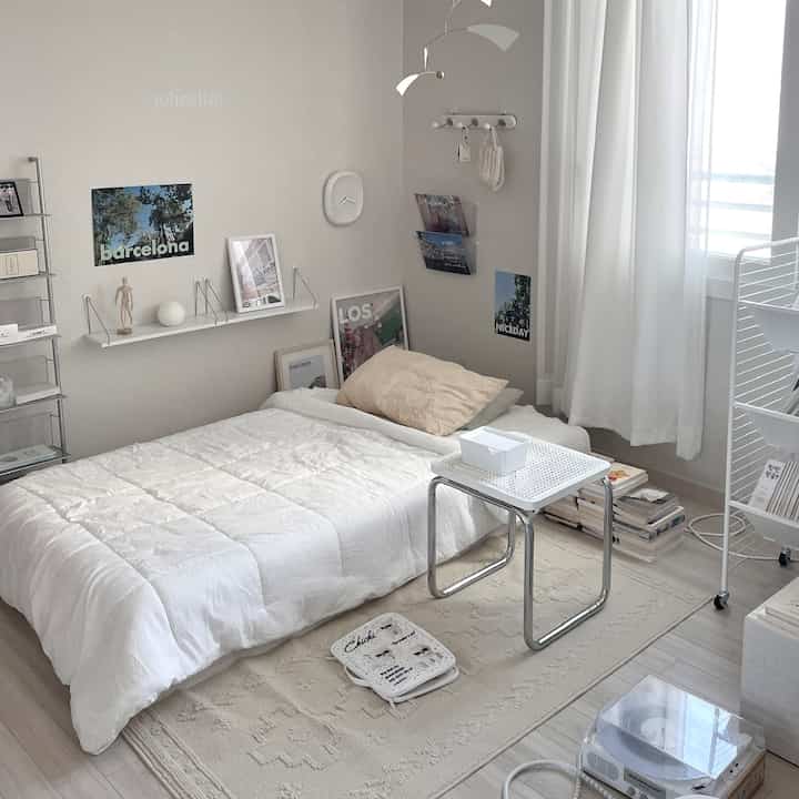 ホワイトとナチュラルトーンの寝室、整った掛け布団とスツール、壁の棚と時計が配置された落ち着いた空間