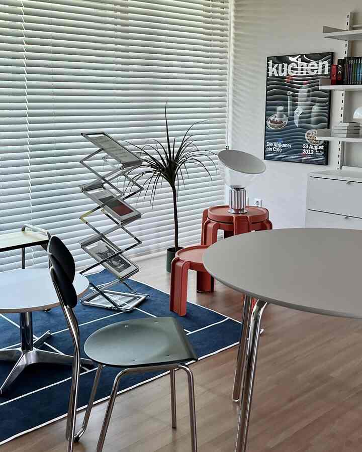 Clean Mid-Century Modern style space with white blinds, blue rug, red stools, bookshelf, and round table