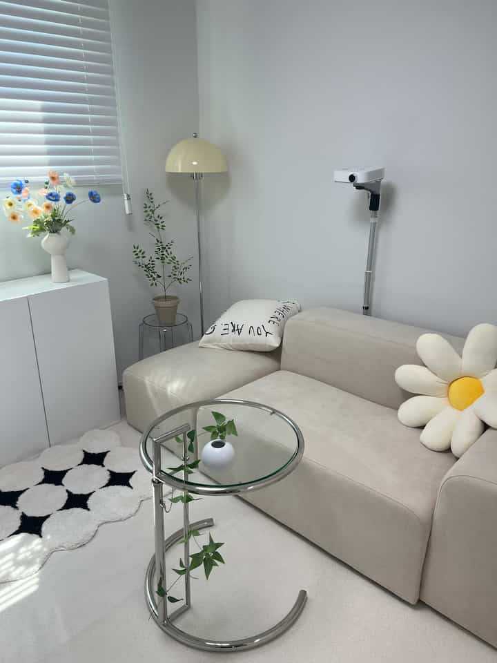 淡いベージュとホワイトトーンの2人掛けソファ、フロアランプ、ガラスのサイドテーブルが配置された自然光あふれるホームゲームと読書の空間