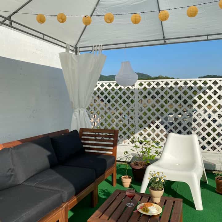 白いテントと緑の人工芝が敷かれた屋上テラス、木製ソファとアウトドアチェア、ストリングライトが配置された雰囲気のあるペット向け空間