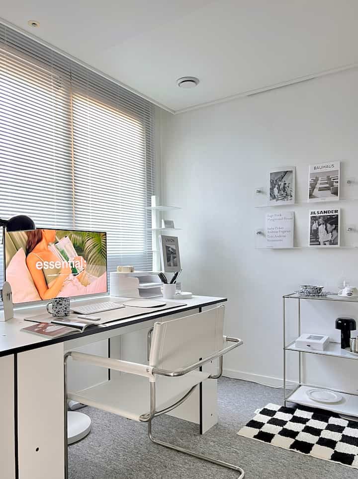 White-toned studio home office with a clean desk and chair, monitor, shelves, and black-and-white checkered rug in a simple modern interior