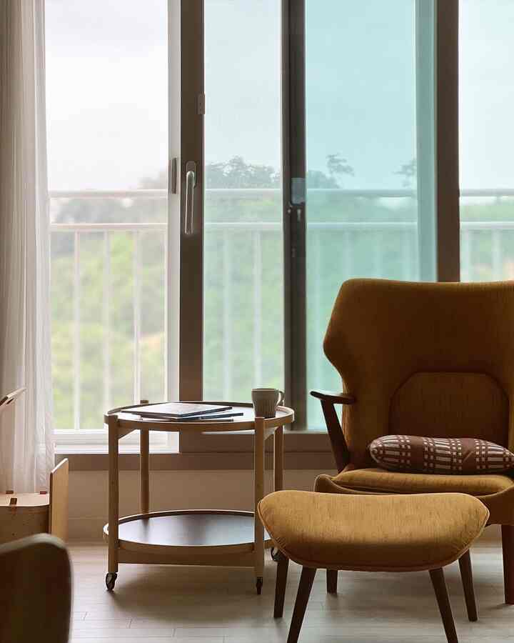 ウッドトーンとマスタード色の組み合わせが印象的なリビング、窓際に配置されたアームチェアとオットマンがある居心地の良い空間