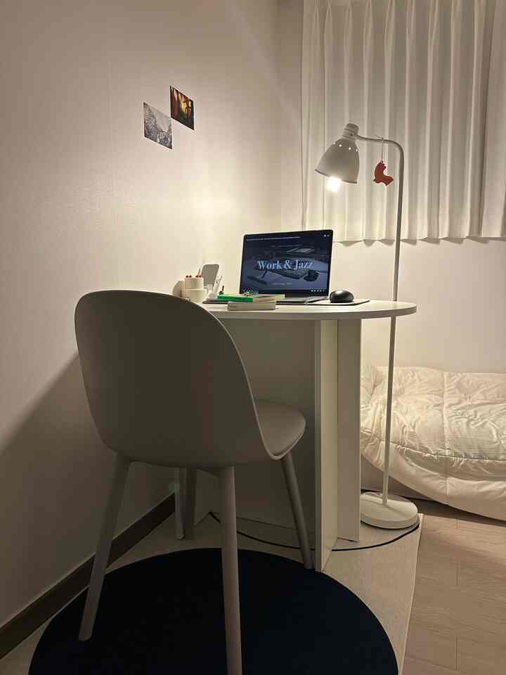 白を基調とした小さな寝室兼ホームオフィス、デスクにノートパソコンとスタンドライト、シンプルな家具が配置された雰囲気のあるインテリア