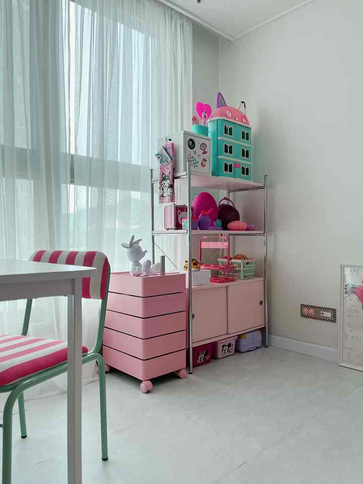 白い壁とカーテンの子供部屋、ピンクの収納棚とトロリーが配置され、おもちゃでかわいらしい雰囲気のインテリア