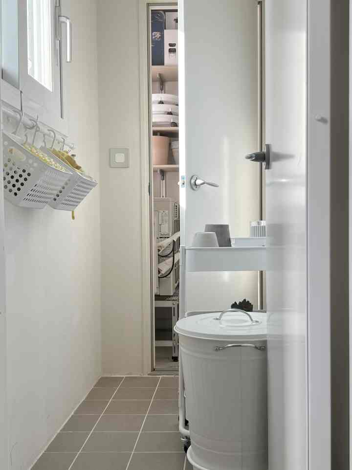 Bright white and gray toned narrow vertical space featuring a 3-tier rolling trolley, round trash can, and window showcasing practical space usage