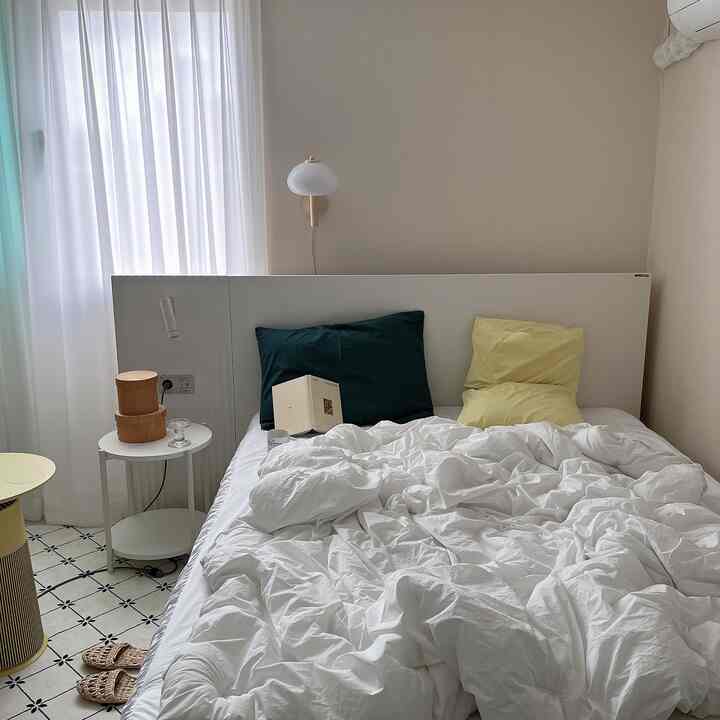 Natural tone beige-walled bedroom with white bedding, a nightstand featuring storage boxes, and sheer curtains creating a simple atmosphere