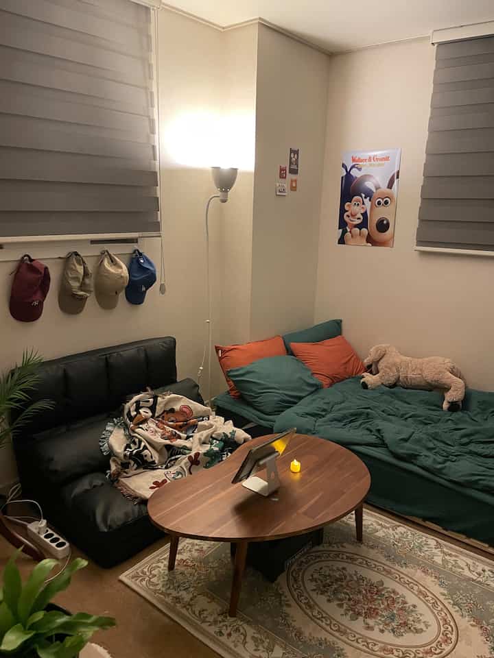 ブラウン系の木目調とグリーンの寝具が調和した4.5畳のワンルーム、コンパクトなソファと照明が配置された居心地の良い休息空間