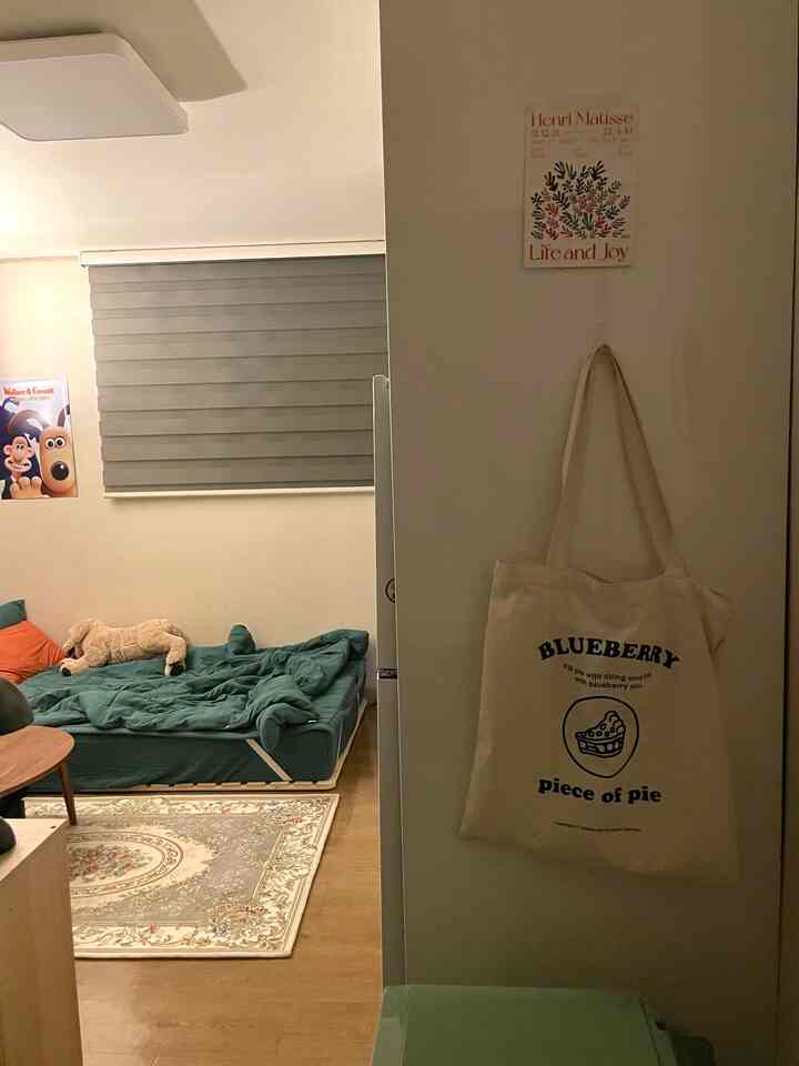 ベージュ色の壁と木目調の床がある玄関から見えるシンプルで温かみのある寝室空間、ポスターとエコバッグが飾られている