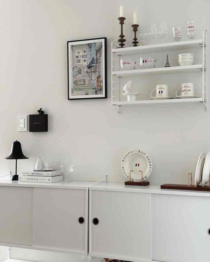 ホワイトトーンのキッチンスペース、壁に設置されたストリングシステム棚とすっきりした収納キャビネットが配置されたミニマルなインテリア