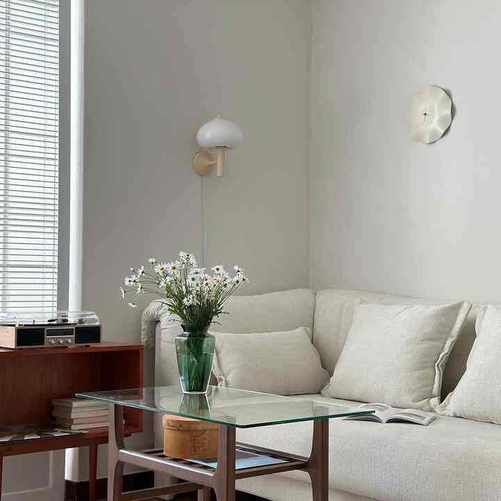 柔らかい白色のリビング、曲面の壁掛け時計とウォールランプ、木製とガラスのコーヒーテーブルに植物の装飾が配置されたシンプルで自然な雰囲気のインテリア