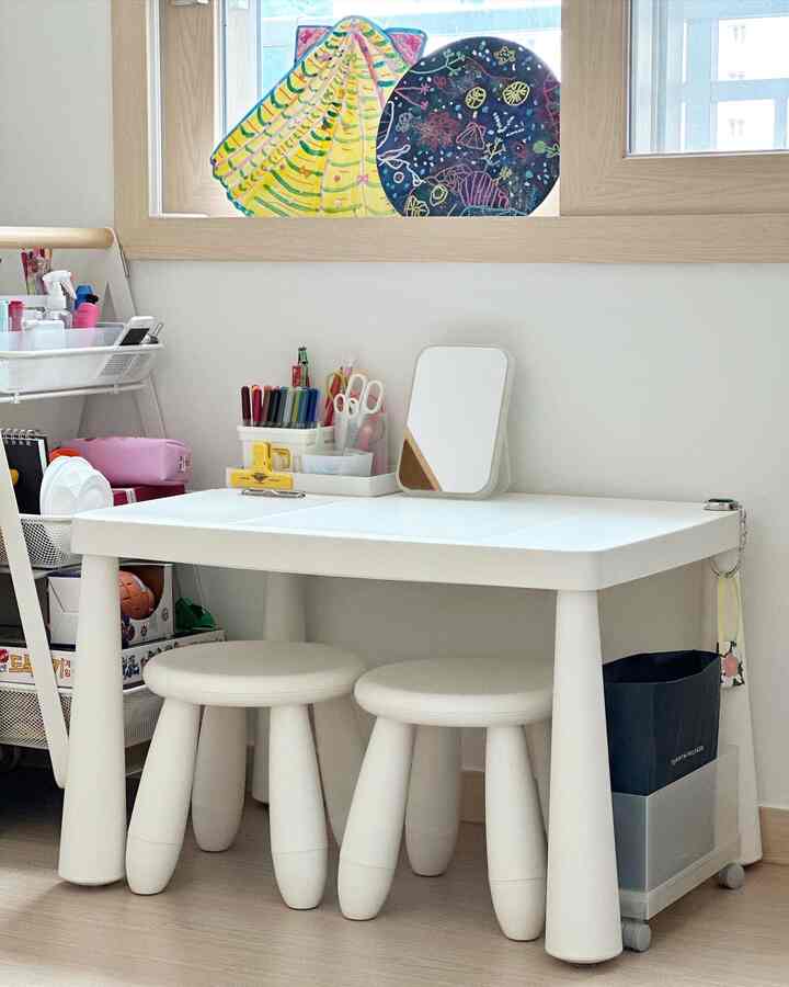 Bright white-toned kids' room featuring a white kids' desk with three stools, organized art supplies, and a storage cart below the window creating a neat interior