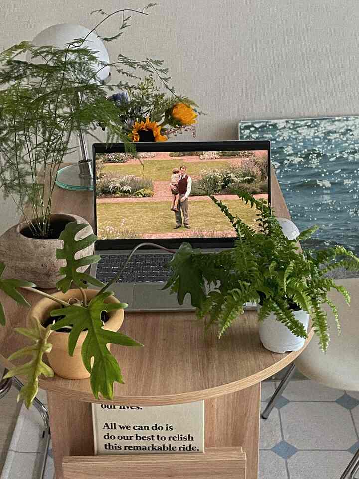 Green and ivory toned studio apartment featuring a wooden round table with plants and a laptop, creating a cozy atmosphere