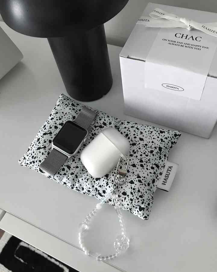 モノトーンのテーブルに黒いテーブルランプと白い箱、斑点模様のクッションの上に時計とエアポッズが置かれた、整然とした小物収納スペース