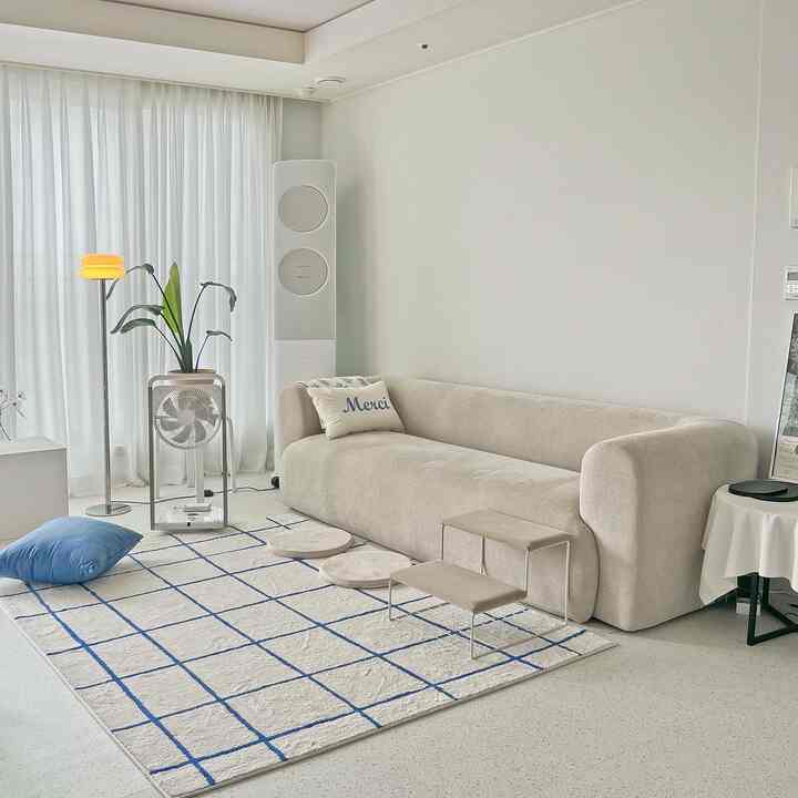 Beige and white toned living room featuring large sofa, blue grid rug, pet steps, and white curtains for a cool summer atmosphere