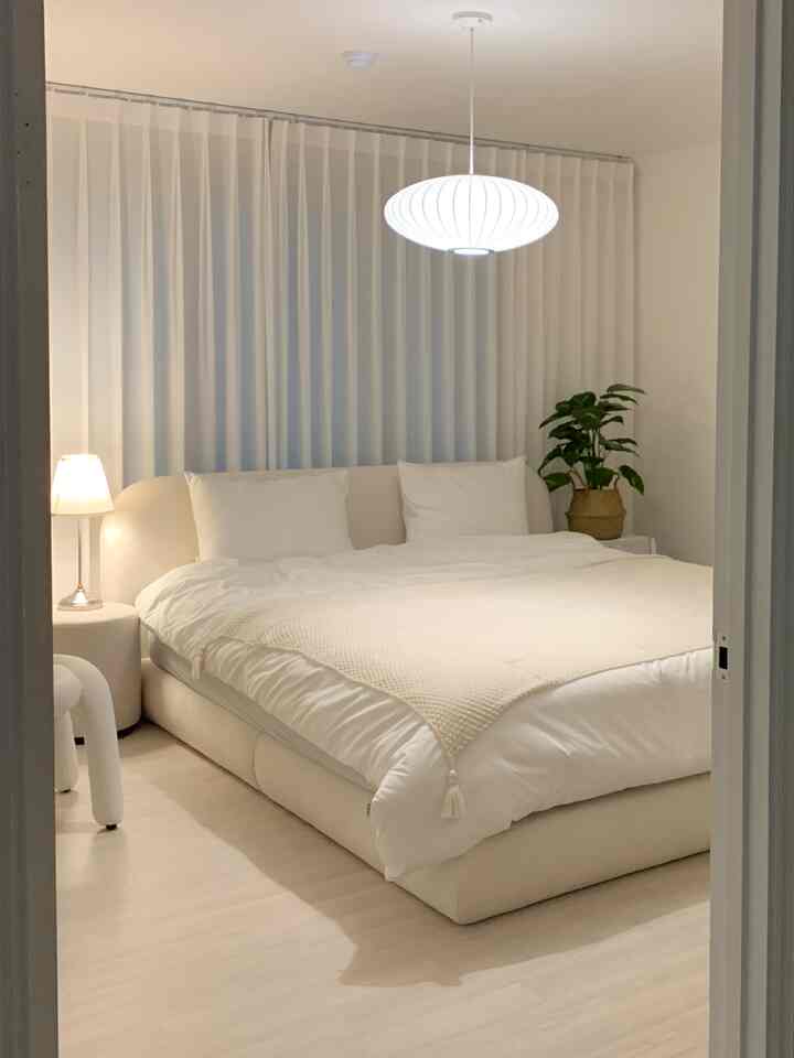 柔らかなホワイトトーンの寝室、中央に大きなベッドとペンダントライト、左側にスツールとナイトテーブル、右隅に植物が配置された自然な雰囲気のあるインテリア