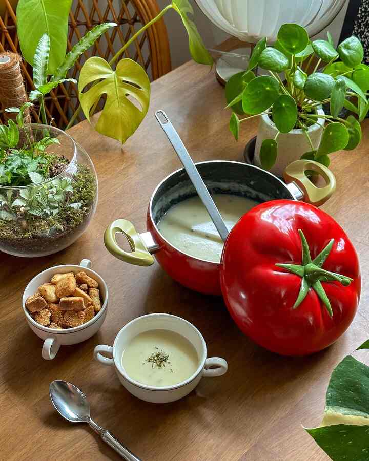 木目調のダイニングテーブルに赤いトマト型の鍋とクリーミースープ、多様な植物が配置されたナチュラルな空間