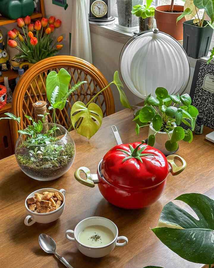 温かみのある木目調のダイニングテーブルに赤いトマト鍋と緑豊かな植物が配置された雰囲気のある空間
