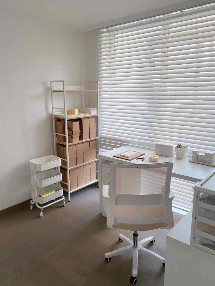 ホワイトとベージュのトーンでまとめられたミニマルなワークスペース、デスクとオフィスチェア、整理された棚とブラインドが配置された清潔感のある空間