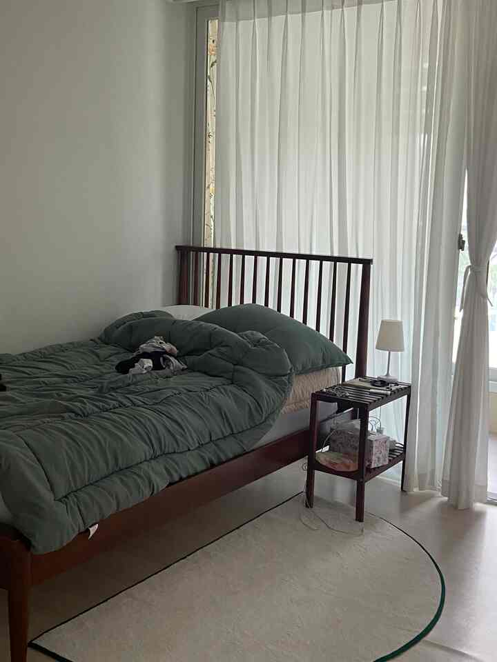Bright white and brown toned bedroom featuring wooden bed and nightstand, green bedding, and white curtains creating a cozy atmosphere
