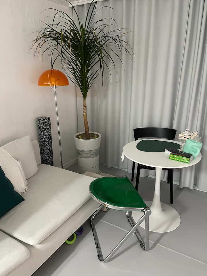 Small space in white and gray tones featuring a minimal sofa, round dining table, green stool, and a large potted plant for a clean living room ambiance