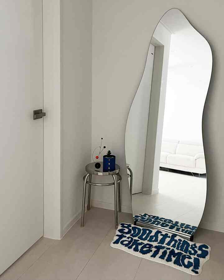 白とシルバーのトーンの玄関フォトゾーン、不規則形の全身鏡とステンレス製の丸椅子、青い文字のラグが配置されたシンプルでモダンなインテリア