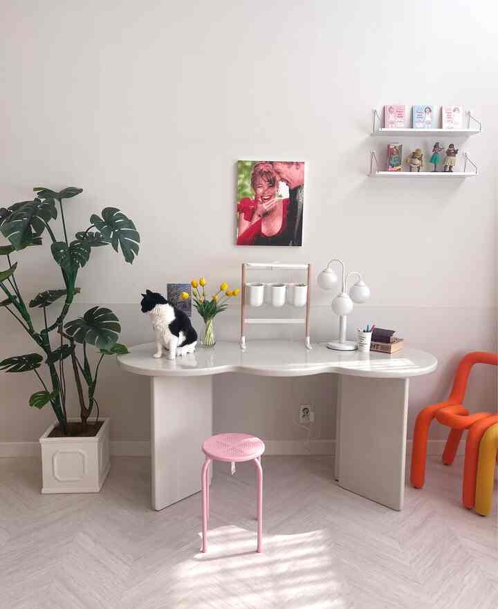 White and green toned dining room with kidney-shaped table, stool, artificial monstera plant, and a black-and-white cat creating a warm modern atmosphere