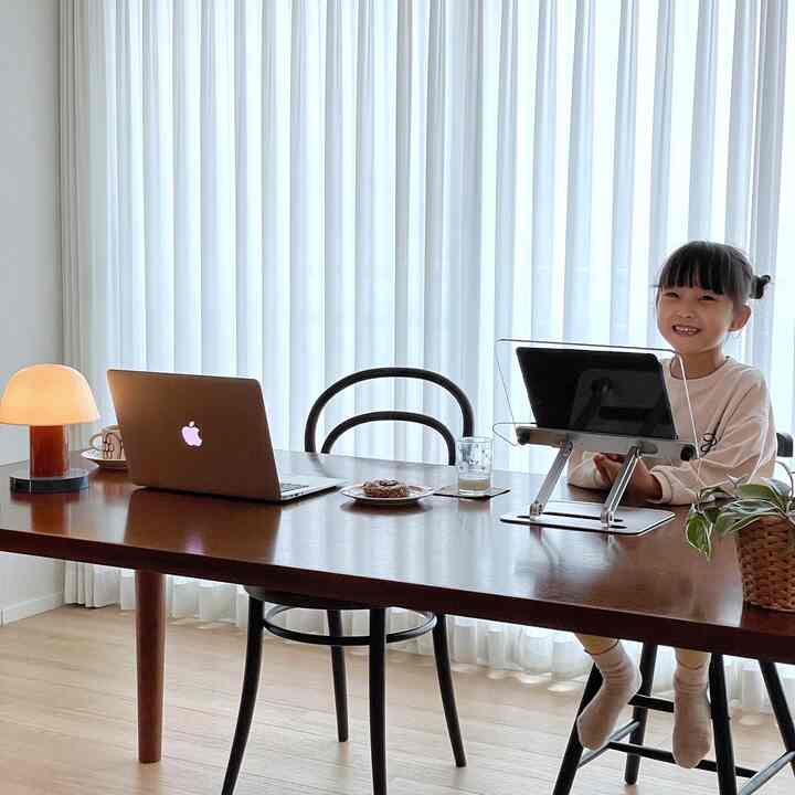 ナチュラルな木目と白いカーテンを背景にしたダイニングテーブルと2脚の椅子、子どもが座って笑顔を見せる明るいホームオフィス空間