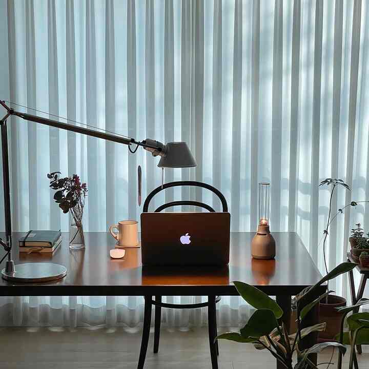 白いシフォンカーテンを背景に、木製のテーブルにアルテミデのテーブルランプ、ノートパソコン、ソニーのブルートゥーススピーカーが配置されたホームオフィスの空間