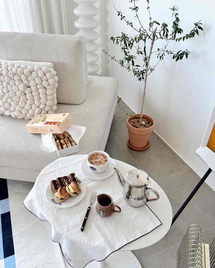 Natural and white toned living room featuring a cream sofa, round coffee table with coffee and desserts, potted plant, and floor lamp creating a cozy atmosphere