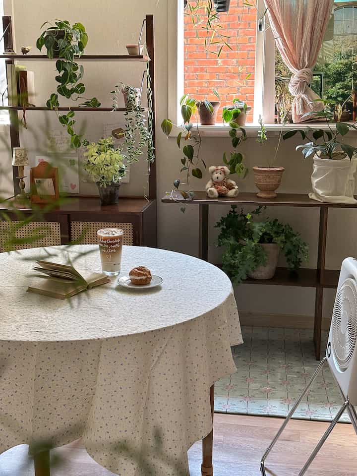 柔らかなベージュ色調のリビング、木製の棚に植物が配置され、丸テーブルにホームカフェの飲み物が置かれた雰囲気のあるインテリア
