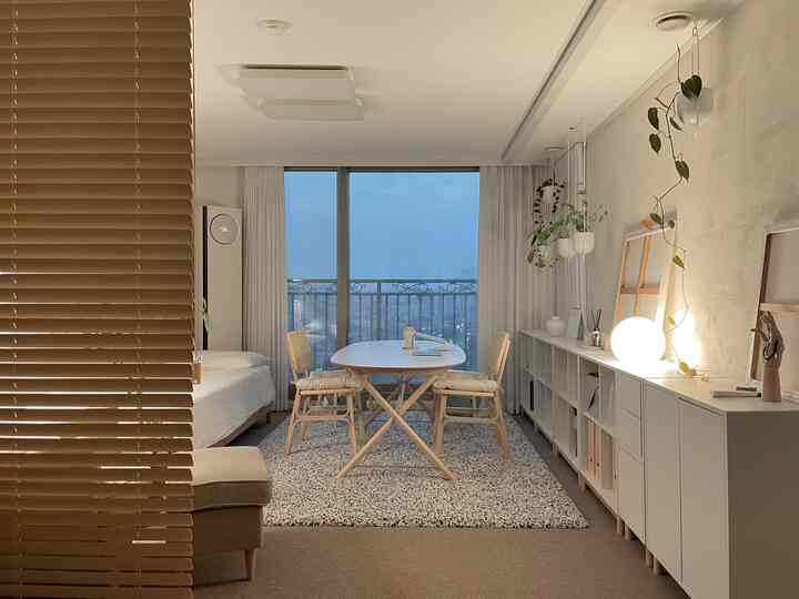 柔らかいベージュとホワイトの色調のリビング、中央にダイニングテーブルと椅子が配置され、植物と棚が壁面にある居心地の良いインテリア