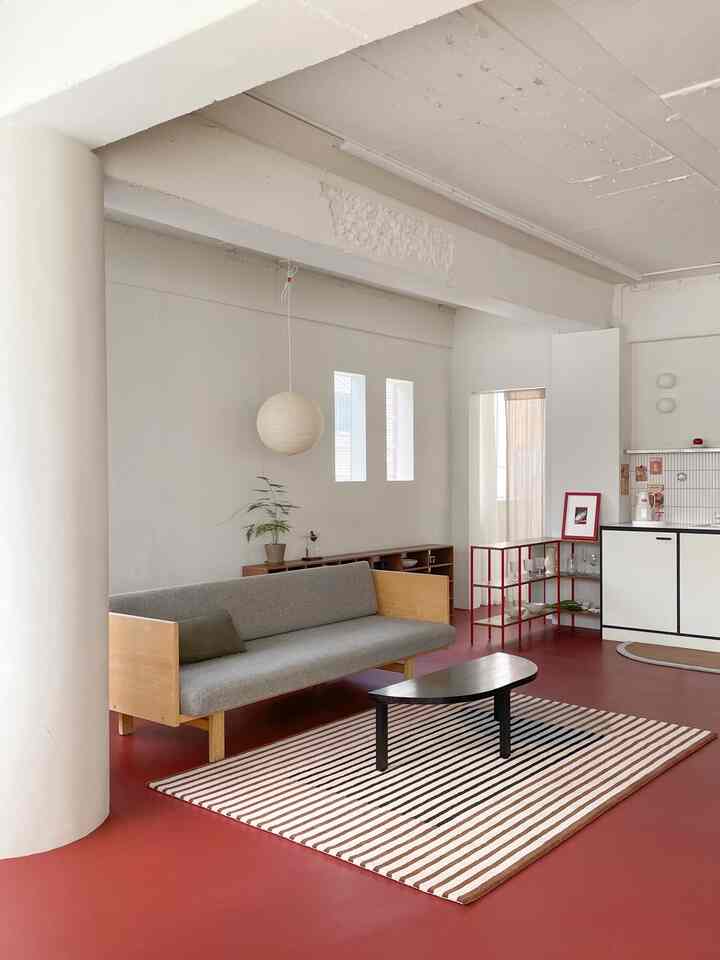 A modern living room with light white walls and red flooring, featuring a natural wood sofa and striped rug
