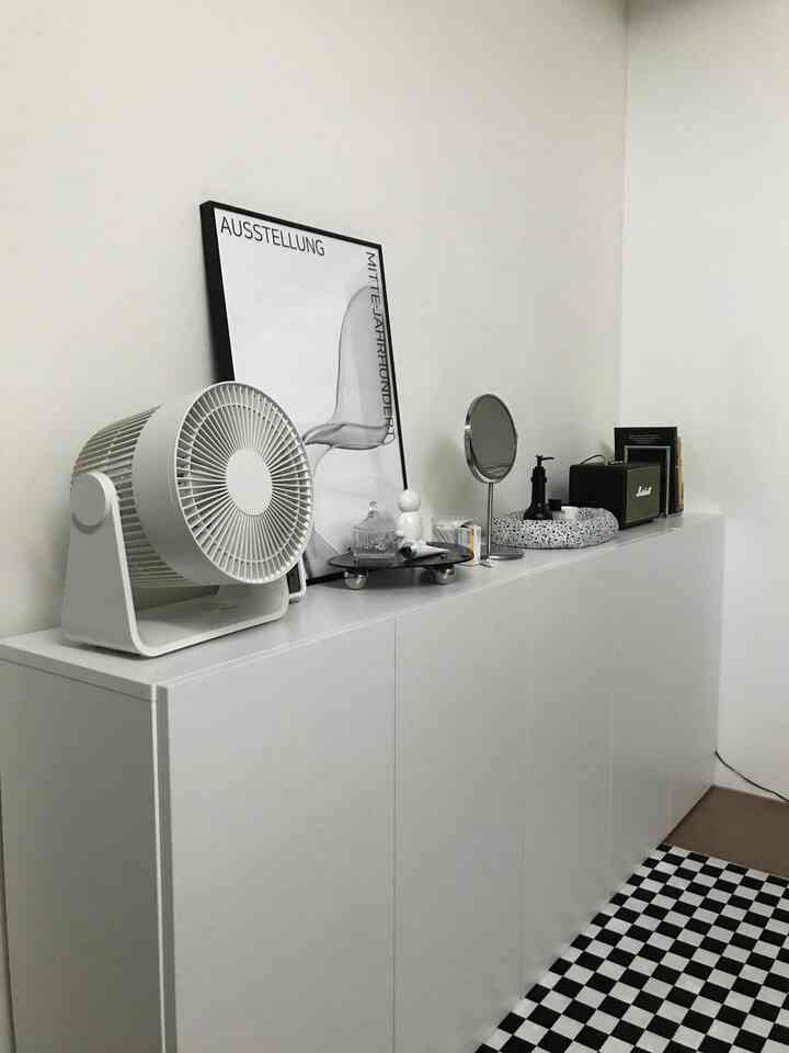 白と黒のモノトーンが印象的なリビングルーム、白いキャビネット上にポスター、卓上扇風機、鏡が配置されたモダンな空間