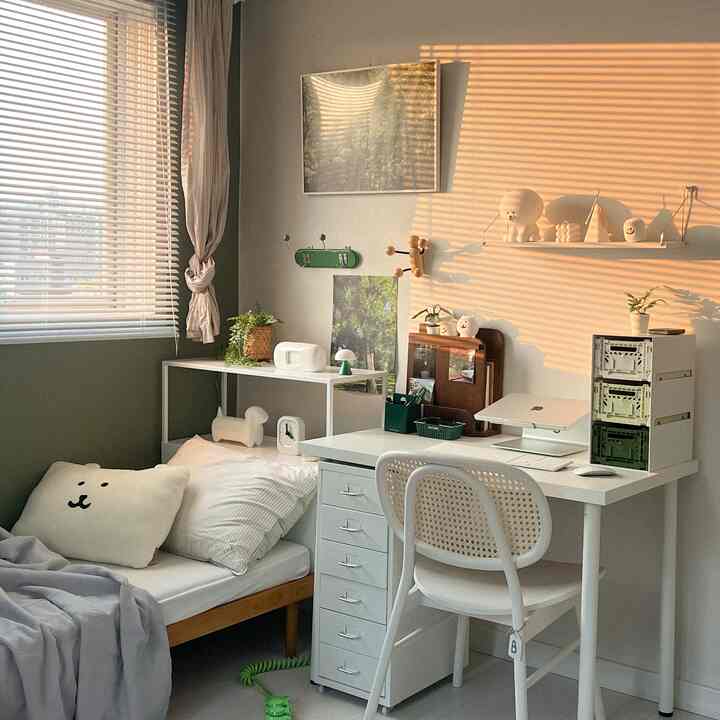 Warm sunset-lit white and green bedroom home office space featuring blinds, tidy desk, and a nature poster