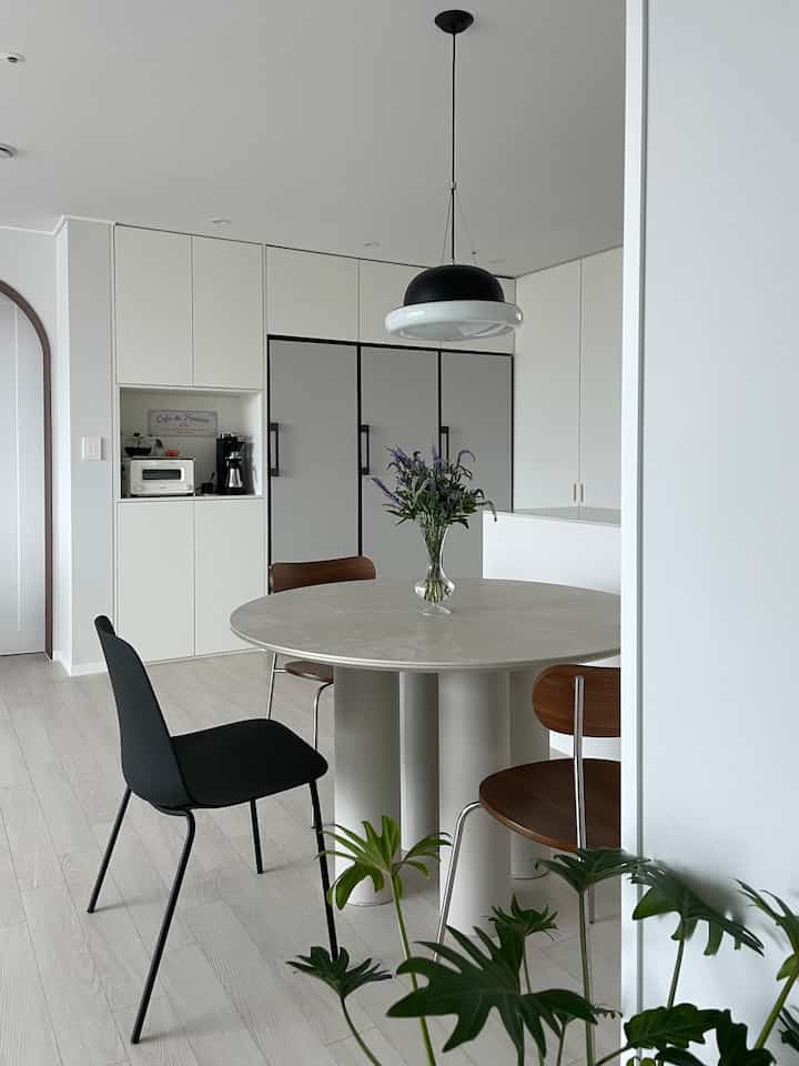 白を基調としたキッチンに黒い円形ペンダントライト、丸いダイニングテーブルと木製チェアが配置されたモダンでナチュラルな空間
